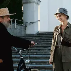 Scena z filmu Jacka Bromskiego: mężczyzna w słomkowym kapeluszu i czarnej sutannie przy rowerze rozmawia z kobietą w wojskowym kapeluszu.