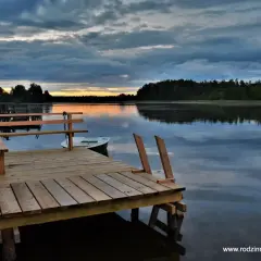 Drewniany pomost nad spokojnym jeziorem Chańcza, z łódką przycumowaną do brzegu. Zachód słońca maluje niebo na pomarańczowo.