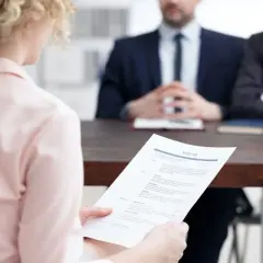 Kobieta z kręconymi włosami trzyma CV podczas rozmowy rekrutacyjnej. Rekruterzy siedzą naprzeciwko.