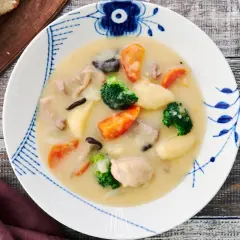 A hearty bowl of Japanese cream stew, brimming with chicken, carrots, potatoes, broccoli, and mushrooms, served with crusty bread.