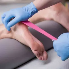 Fizjoterapeuta zakłada taśmę kinezjotaping na stopę, wspierając ćwiczenia na ostrogę piętową.