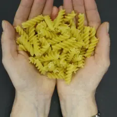 Dłonie trzymające porcję żółtego makaronu fusilli na ciemnym tle.