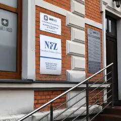 Elewacja budynku z cegły z tabliczkami "Santi Med" i "NFZ", drewnianym oknem i drzwiami.