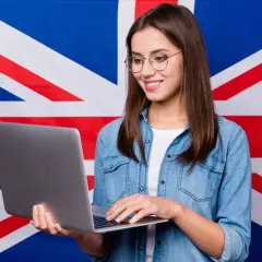 Uśmiechnięta dziewczyna w okularach pracuje na laptopie na tle flagi Wielkiej Brytanii, tworząc przykładowy artykuł po angielsku.