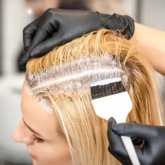 Fryzjerka w czarnych rękawiczkach nakłada farbę na blond włosy, wykonując farbowanie odrostów.