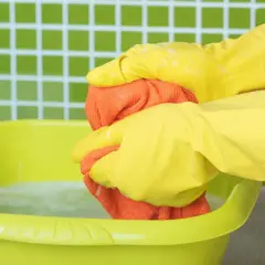 Ręce w żółtych rękawiczkach wyciskają pomarańczową ściereczkę nad zieloną miską z wodą. Dowiedz się, jak usunąć żywicę z ubrania.
