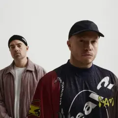 Dwóch mężczyzn w czapkach, jeden w beżowej kurtce, drugi w kolorowej bluzie. Wyglądają jak zespół problem, gotowi na wyzwania.