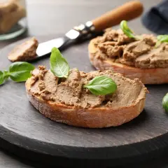 Dwa kawałki chleba z pyszną, domową pastą z mięsa z rosołu, ozdobione listkami bazylii, na drewnianej desce.