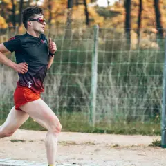 Mężczyzna w sportowym stroju biegnie, ćwicząc jak zwiekszyc kadencje w bieganiu. W tle siatka i drzewa.