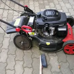 Kosiarka z czerwonymi detalami i solidnymi kółkami do kosiarki, gotowa do pracy na podjeździe z kostki brukowej. Obok leży miotka.