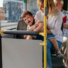 Płaczące dziecko na rękach matki w autobusie, obok siedzi starszy mężczyzna.