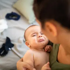 Un bebé con cara de llanto es sostenido por su madre. Parece que el bebe se cae de la cama y está asustado.