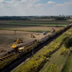 Długi pociąg z wagonami wypełnionymi ziemią, obok koparki i samochodów. Prace torowe w tle pól i zabudowań, widać odbojnica kolejowa.