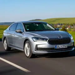 Srebrna, nowa Skoda Superb pędzi po drodze, mijając malownicze, zielone wzgórza i domki.