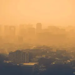 Vista aérea de una ciudad envuelta en una densa neblina anaranjada, con siluetas de edificios apenas visibles. Las partículas PM10 flotan en el aire, creando esta atmósfera opresiva.