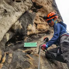 Młoda osoba w kasku wspina się po skale na via ferrata w Decinie. Tabliczka "Cesta do nebíčka" wskazuje trudność trasy.