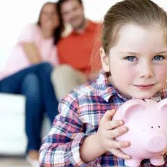 Dziewczynka z różową świnką skarbonką. Rodzice w tle. Czy komornik może zająć konto dziecka?