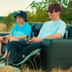 Dwóch chłopców gra w gry wideo na czarnej kanapie na łonie natury. Jeden z nich, młodszy, ma na głowie czarną czapkę typu bucket hat, a drugi, starszy, nosi okulary przeciwsłoneczne. Wyglądają jakby byli na planie teledysku rapera.