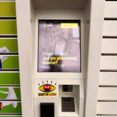 Ekran automatu InPost z uśmiechniętym mężczyzną i pytaniem "Dzień dobry! Już po pierwszej kawie?". Obok widać produkty z półek.