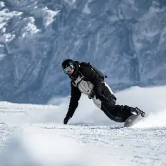 Snowboarder w kasku i goglach, z ochraniaczem nadgarstka snowboard, pochyla się nisko, tworząc obłok śniegu podczas zjazdu.