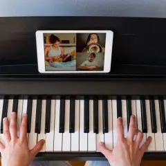 Ręce na klawiaturze pianina cyfrowego, na ekranie tabletu widać lekcję muzyki online z uczniami grającymi na ukulele, trąbce i gitarze.