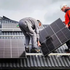 Deux techniciens travaillent sur une toiture pour l'installation de panneaux solaires. L'un tient un panneau, l'autre est penché.