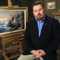 A man in a suit sits beside a Thomas Kinkade painting of a lighthouse on a cliff.