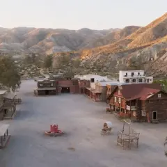 Un pueblo del oeste con edificios de madera y un pozo de agua, rodeado de colinas áridas.
