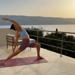 Mujer en postura de yoga en terraza con vista al mar al atardecer. Un momento de **slow life** y conexión.
