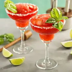 Dwa mrożone strawberry daiquiri z bazylią i cukrowym brzegiem, ozdobione plasterkami truskawek. Obok leżą ćwiartki limonki.