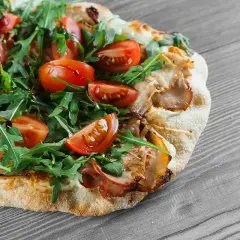 Une délicieuse pinsa recette garnie de tomates cerises, roquette fraîche et viande effilochée, sur fond de bois gris.