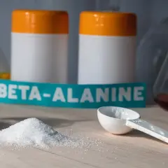 Biały proszek beta-alaniny na blacie, obok miarki i słoiczka. W tle dwa białe pojemniki z pomarańczowymi nakrętkami. Czy beta-alanina skutki uboczne są powodem do obaw?