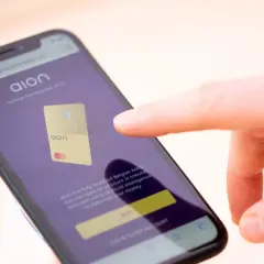 Palec dotyka ekranu telefonu z aplikacją Aion bank, prezentującą złoty debetowy Aion.