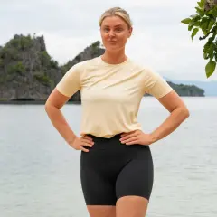 En kvinna med blont hår står på en strand med händerna på höfterna. Bakom henne syns klippor och hav. Hon ser ut som en robinsonvinnare.