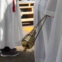 Osoba w białej szacie trzyma złote kadzidło na łańcuszku.