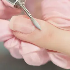 Dłoń w różowej rękawiczce usuwa frezarką resztki lakieru hybrydowego z paznokcia.