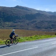 A lone cyclist enjoys a scenic bike tour Scotland, with rolling hills and mountains in the background under a bright blue sky.