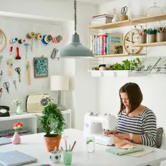 Mujer cosiendo en su **taller de manualidades en casa**. Pared con herramientas, estanterías con libros y plantas.