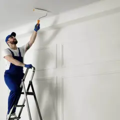 Mężczyzna w niebieskim kombinezonie na drabinie maluje sufit wałkiem. Pokazuje, jak malować sufit, by uzyskać idealnie gładką powierzchnię.