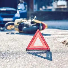 Trójkąt ostrzegawczy na drodze po wypadku. Złoty skuter leży na boku, obok niebieski samochód z otwartą maską. Może to być symbol problemów z ubezpieczeniem, jak 2 rata OC po terminie.
