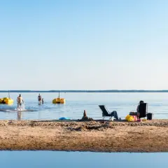 Solig dag vid Karlstad Swecamp Bomstadbaden. Folk badar och kopplar av på stranden med gula trampbåtar i vattnet.