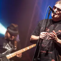 Wokalista Budki Suflera śpiewa na koncercie. W tle basista gra na gitarze. Piosenki Budki Suflera alfabetycznie to ich bogaty repertuar.