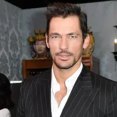 Hombre con traje de raya diplomática, mirada intensa, fondo con candelabro y decoraciones.