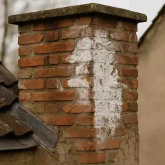 Ziegelkamin mit weißen Ausblühungen, die auf Kondenswasser hindeuten. Daneben ein Ziegeldach und ein verputztes Haus.