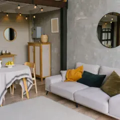 Un salon moderne avec un canapé clair, des coussins colorés et un miroir rond. La table ronde couverte d'une nappe grise et les guirlandes lumineuses ajoutent une touche chaleureuse, reflétant une nouvelle tendance déco.