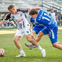 Piłkarze walczą o piłkę na boisku. Jeden w białej koszulce, drugi w niebieskiej z numerem 11. Emocje jak w walce o 3 liga tabele.
