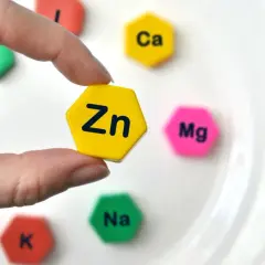 Palec wskazujący trzyma żółty heksagon z napisem "Zn". Wokół niego na białym talerzu leżą inne heksagony z symbolami pierwiastków: Ca, Mg, Na, K.