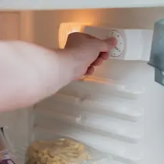 Main d'une personne ajustant le thermostat d'un réfrigérateur, montrant comment fonctionne un frigo. Des aliments sont visibles à l'intérieur.