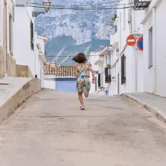 Niña corre por calle empinada de un pueblo de Alicante, con casas blancas y montañas al fondo.