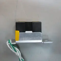 Mały, czarny element z żółtym akcentem i przewodami, przypominający wtyczkę RGB, zamontowany na metalowej płytce.
